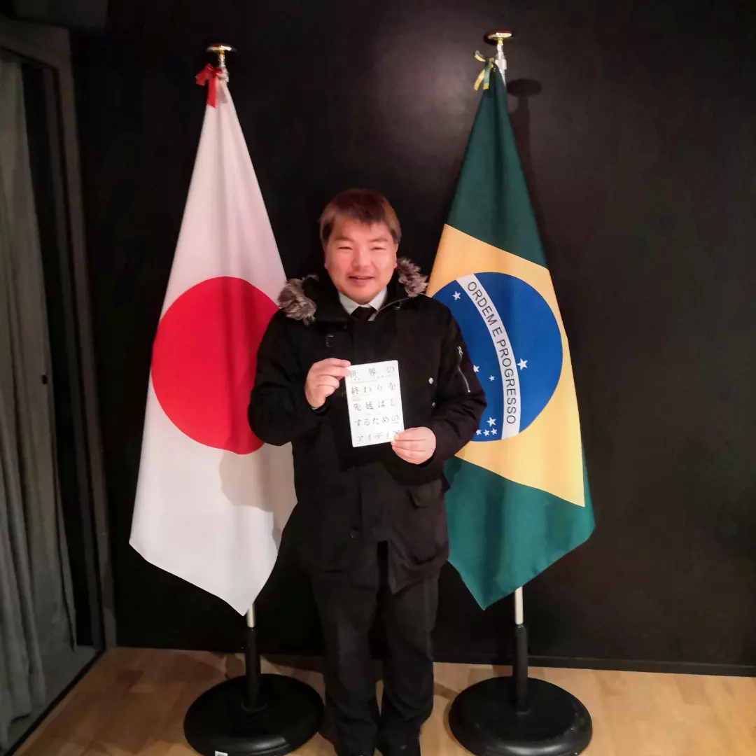 ブラジル国旗と日本国旗の前で『世界の終わりを先延ばしするためのアイディア』を持つ榊原平