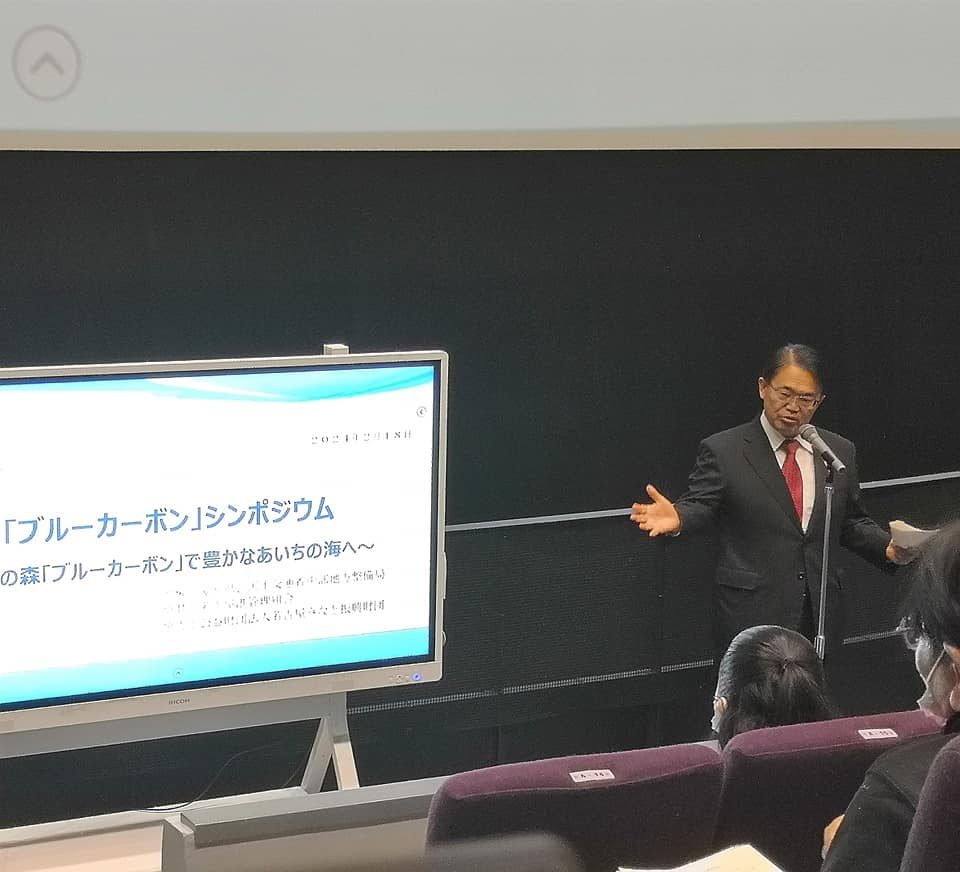 ブルーカーボンシンポジウムと表示されたディスプレーと黒いスーツ姿で立つ大村秀章愛知県知事