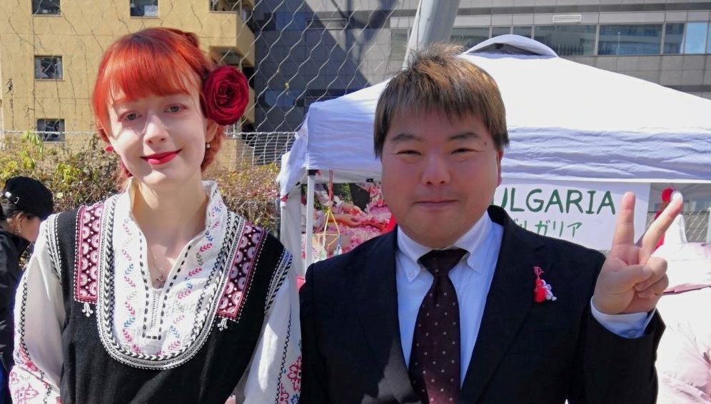 ブルガリア民族衣装を着た赤毛の女性とマルテニツァを身に着け黒いスーツを着た榊原平