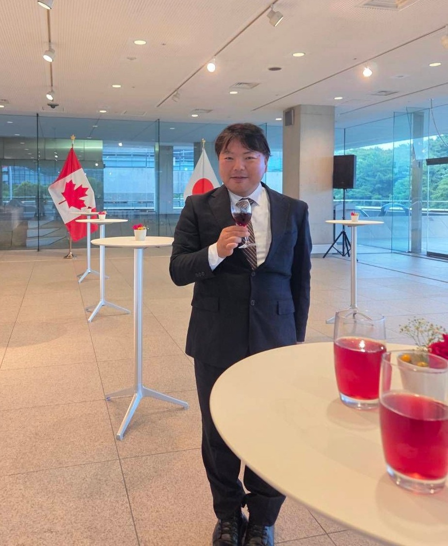 カナダと日本が語る偽情報対策の最前線 3 イベント後の交流会でワイングラスを片手に微笑む筆者、榊原平。背景にはカナダと日本の国旗が見える。