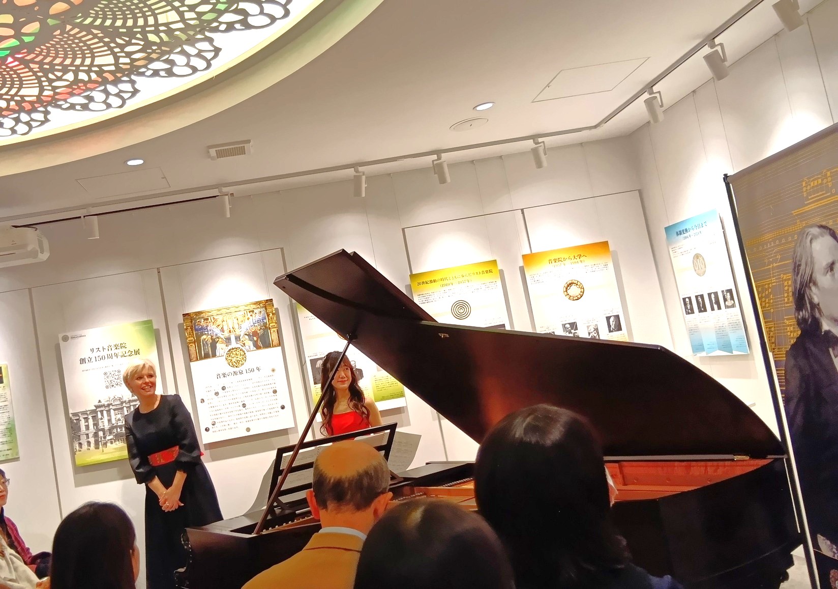 リスト音楽院創立150周年記念展覧会 特別コンサート|天川真奈さんが奏でるベヒシュタインの調べ 2 会場の中心に置かれた艶やかな黒のベヒシュタインのグランドピアノ。赤いドレスを纏った天川真奈さんが演奏し、観客が聞き入っている様子。