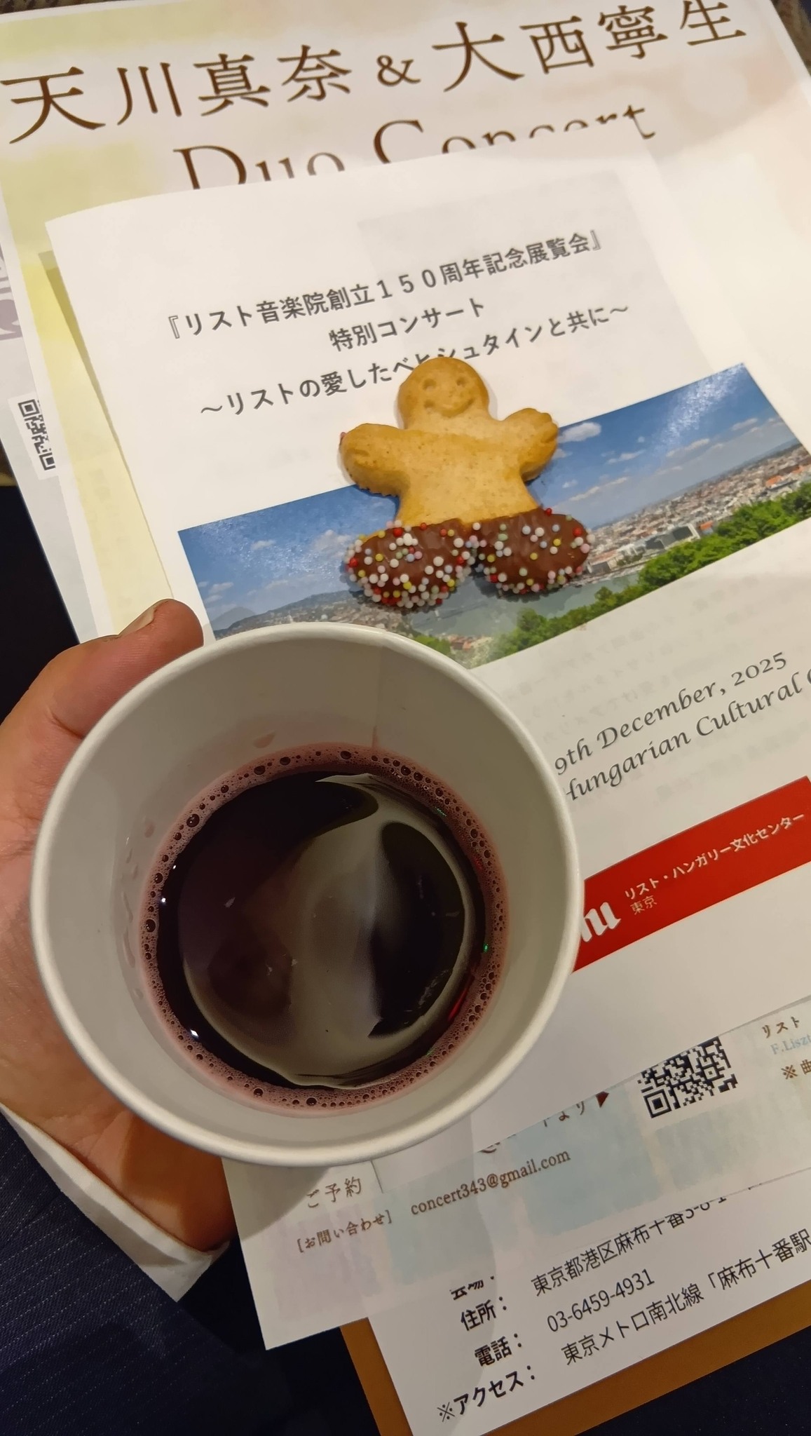 リスト音楽院創立150周年記念展覧会 特別コンサート|天川真奈さんが奏でるベヒシュタインの調べ 1 コンサートのプログラムの上に、赤いホットワインが入った紙コップと、チョコの靴を履いた可愛い人型のジンジャーブレッドが置かれている様子。