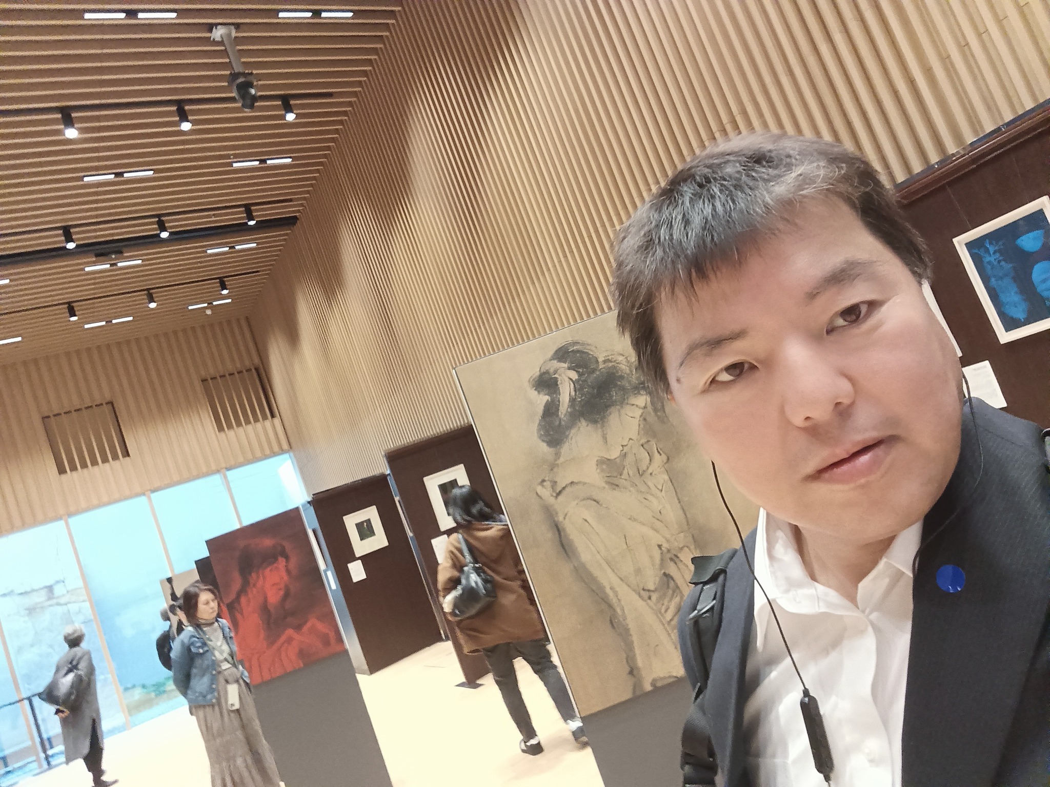 スーツ姿の榊原平氏が「怪談」展の会場内で自撮りをしている。背景は木目調の壁が特徴的な展示空間で、モノクロの人物画や赤い絵画などの作品、他の来場者の姿が見える。