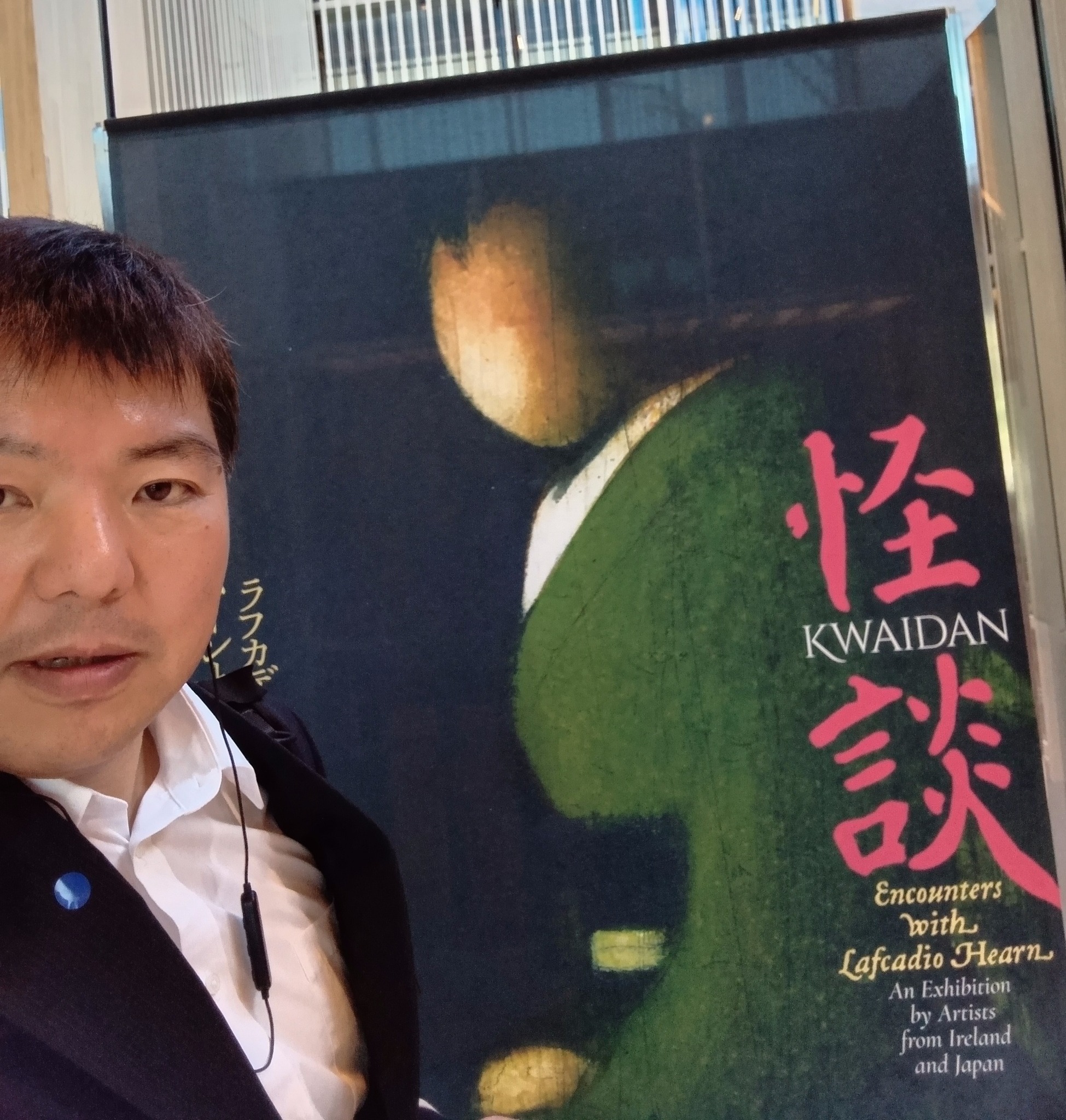 スーツ姿の榊原平氏が「怪談 KWAIDAN」と書かれた展覧会のメインビジュアルポスターの前で自撮りをしている。