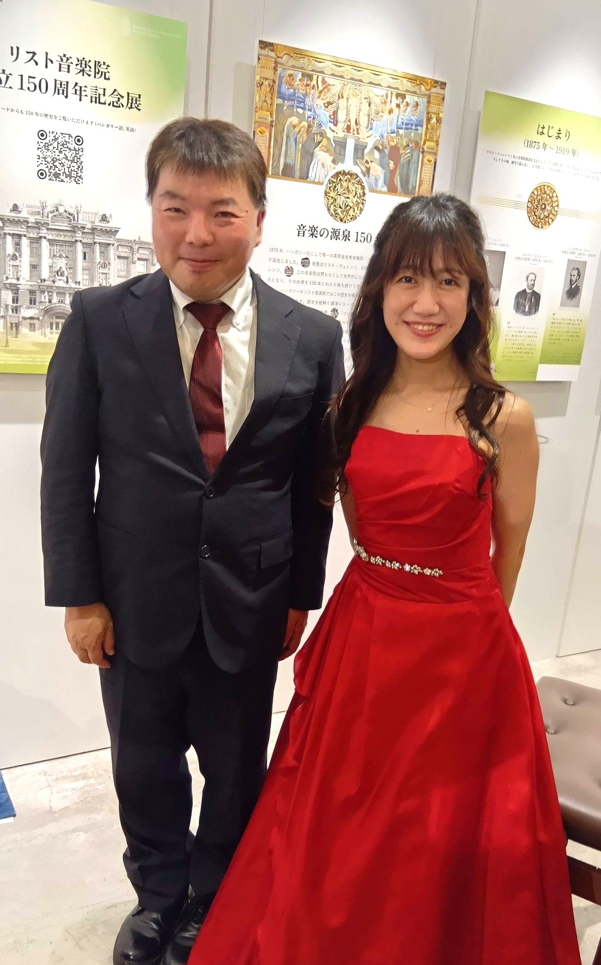 リスト音楽院創立150周年記念展覧会 特別コンサート|天川真奈さんが奏でるベヒシュタインの調べ 4 赤いドレスを着たピアニストの天川真奈さんと、スーツ姿の男性が笑顔で写っているツーショット。