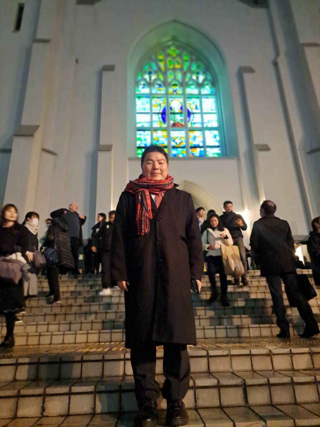 カトリック布池教会の鮮やかなステンドグラスを背に、階段に立つ榊󠄀原平。黒のコートに赤いチェックのマフラーを巻いた冬の装い