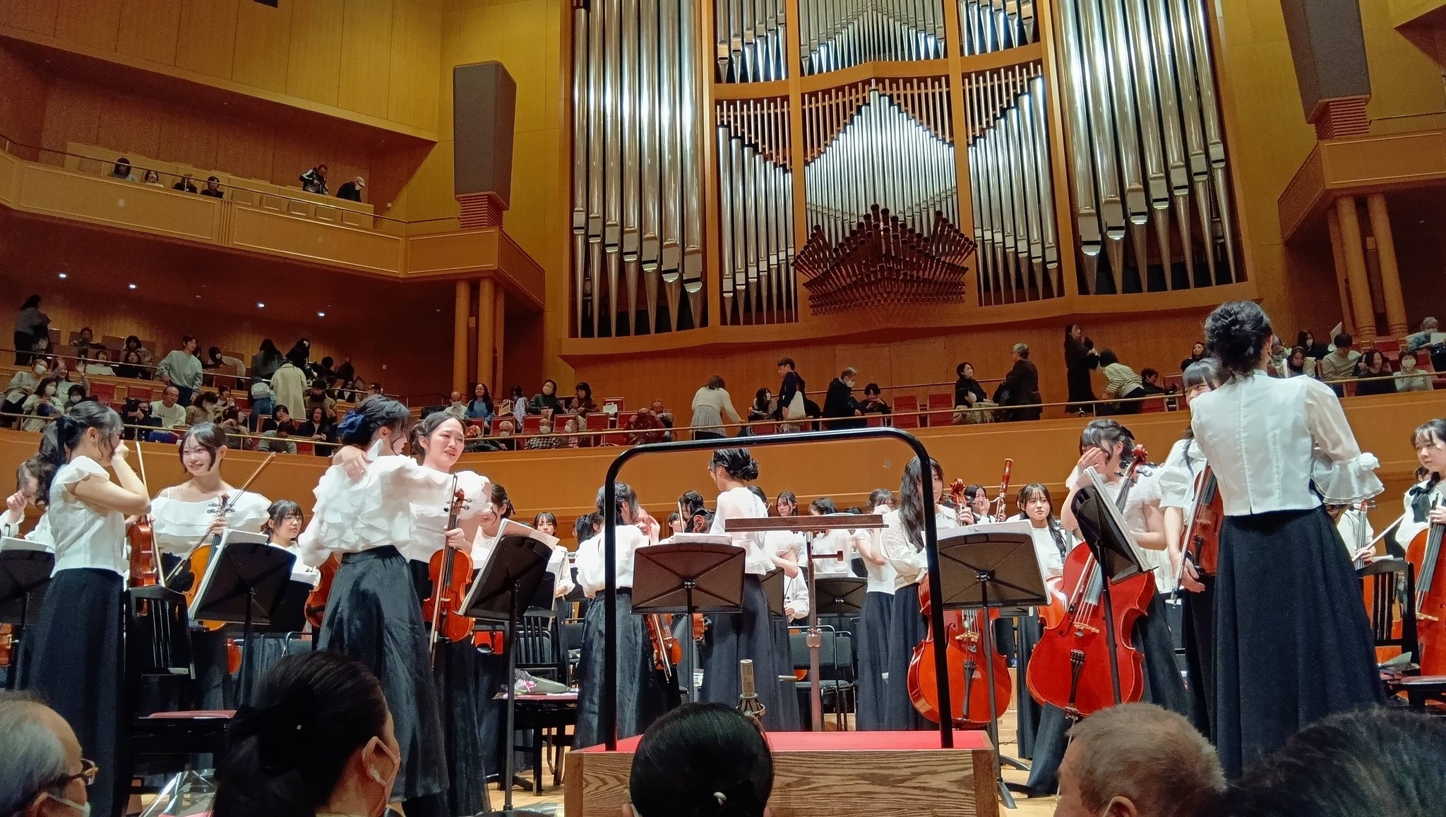 椙山フィルハーモニーオーケストラ 第58回定期演奏会 〜白い翼が響かせた歓喜と涙〜 5 巨大なパイプオルガンがある愛知県芸術劇場コンサートホールの舞台上で、椙山フィルハーモニーオーケストラの第58回定期演奏会を終えた直後、白いブラウスと黒いロングスカート姿の女子生徒たちが感極まって涙ぐみながらお互いに抱き合っている様子。手前には楽器を置いた譜面台や指揮台があり、客席には観客がいる。