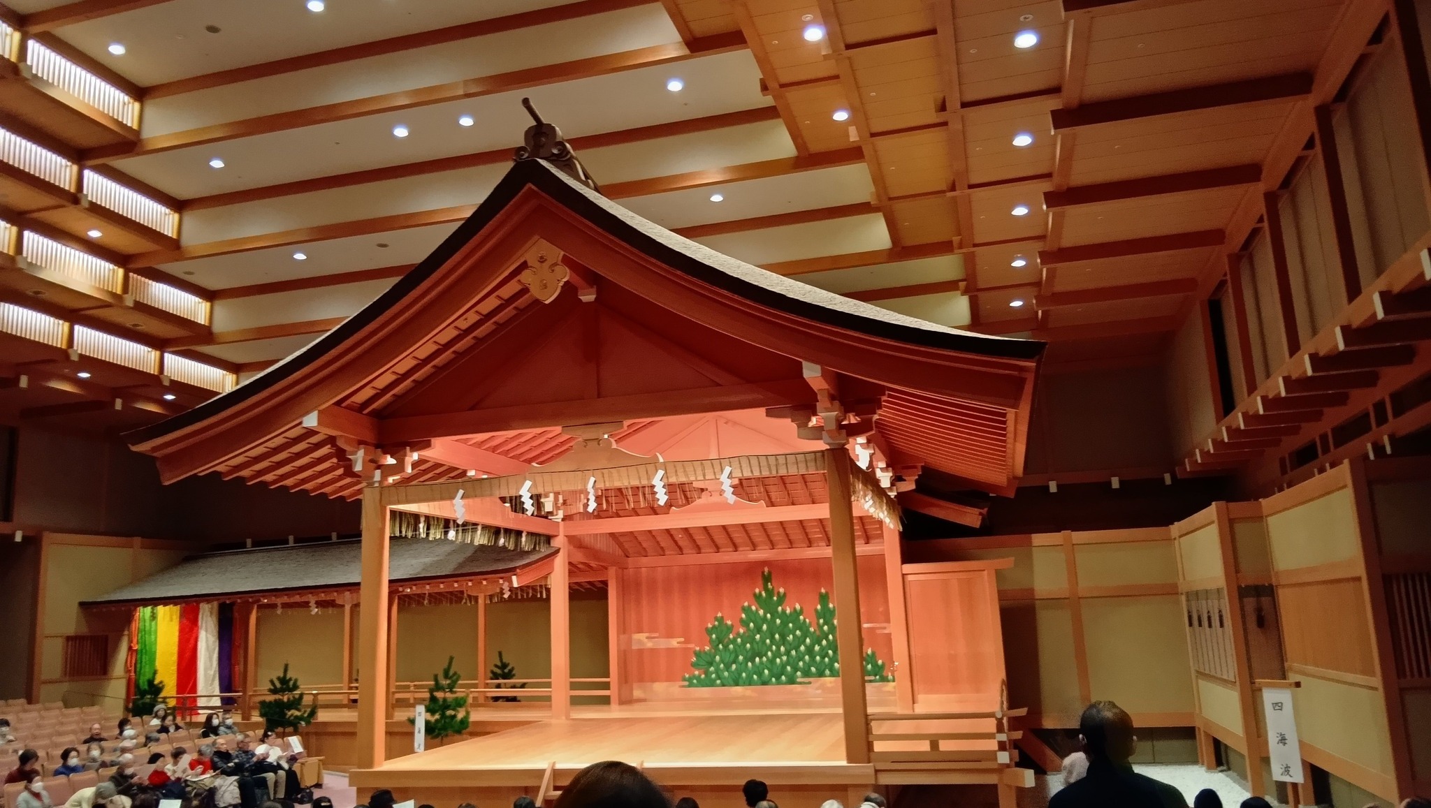 名古屋能楽堂 新春謡初め2026 五流派が揃う感動の伝統芸能 4 名古屋能楽堂の総檜造りの能舞台。背景には緑の松の絵が描かれ、舞台の前には白砂が敷き詰められている。客席の照明が点灯している。