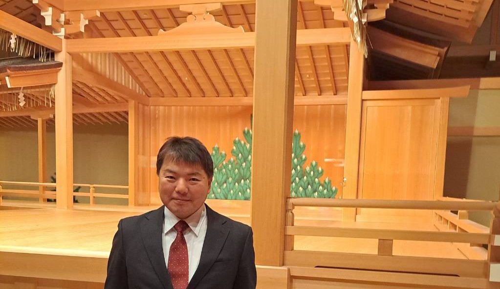 能舞台を背景に立ち、カメラを見て微笑む、黒いスーツに赤いネクタイ姿の榊原平氏の全身写真。