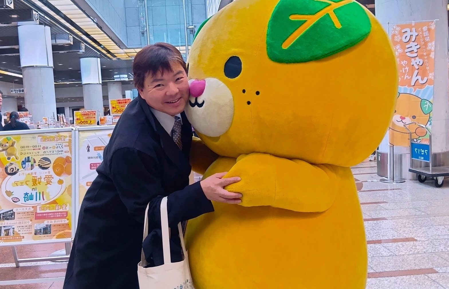 神奈川県の川崎アゼリアで開催された「愛媛フェア in 川崎」にて、愛媛県イメージアップキャラクター「みきゃん」と笑顔で抱き合う榊原平。背景にはイベントののぼりやポスターが写っている。