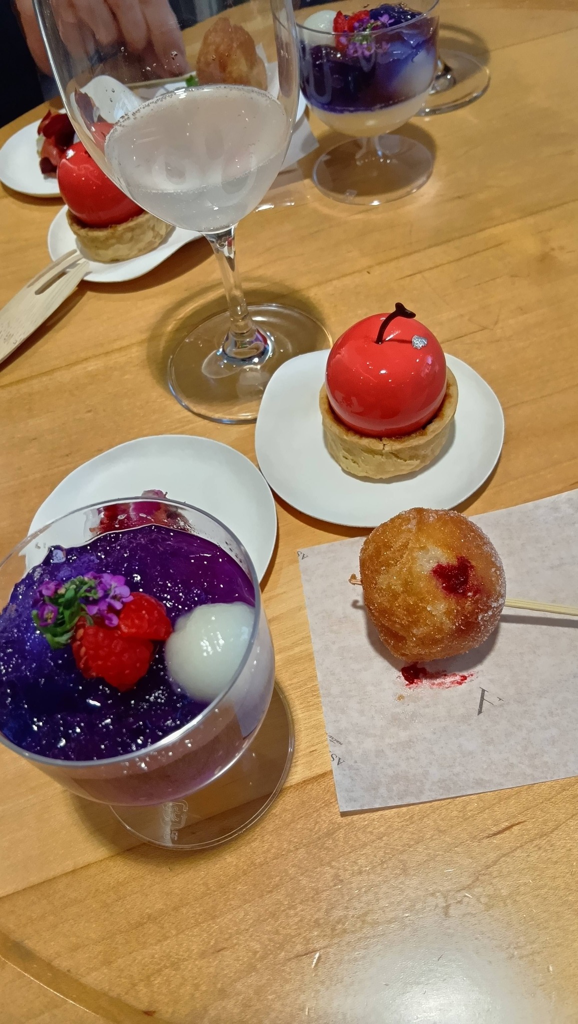 テーブルの上に並べられたデザートの接写。手前には鮮やかな紫色のゼリー、右側にはポーランドの伝統菓子ポンチキ、奥には赤いリンゴの形をしたタルトと飲み物のグラスが配置されている。