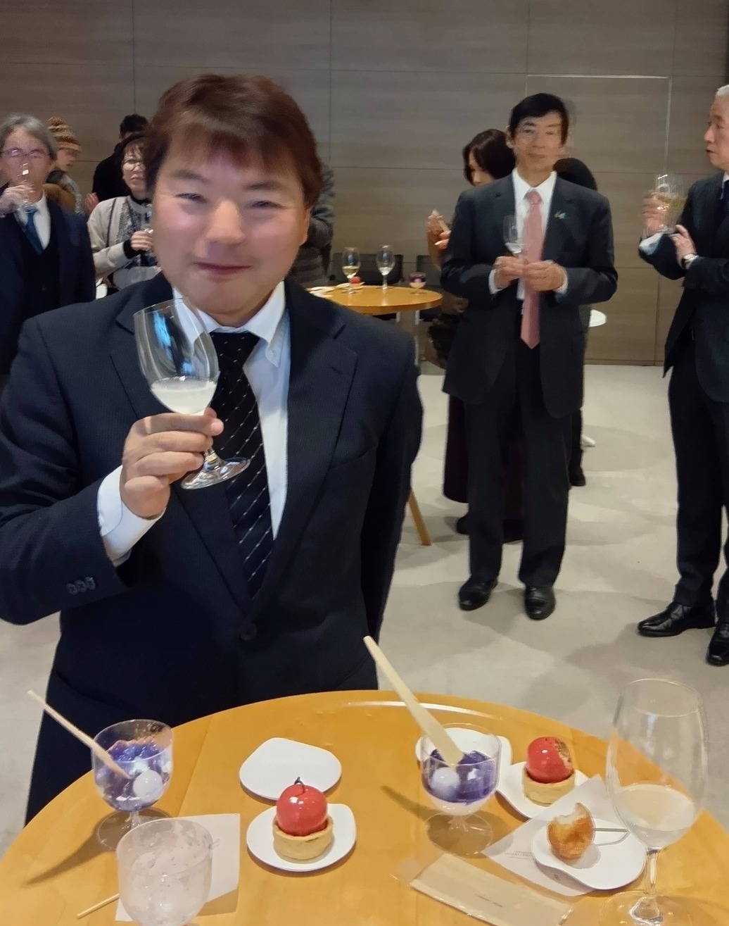 レセプション会場にて。スーツ姿の榊原平氏がグラスを手に持ち、柔らかな表情で立っている。背景には宮島昭夫前駐ポーランド日本大使や他の来場者が談笑している様子が写っている。