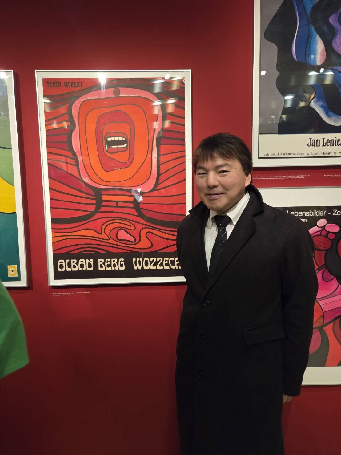 銀座・ギンザ・グラフィック・ギャラリーの展示室内にて。ヤン・レニツァの代表作、オペラ『ヴォツェック』の巨大な赤いポスターの右側に立ち、黒いコート姿で微笑む榊原平氏のポートレート。