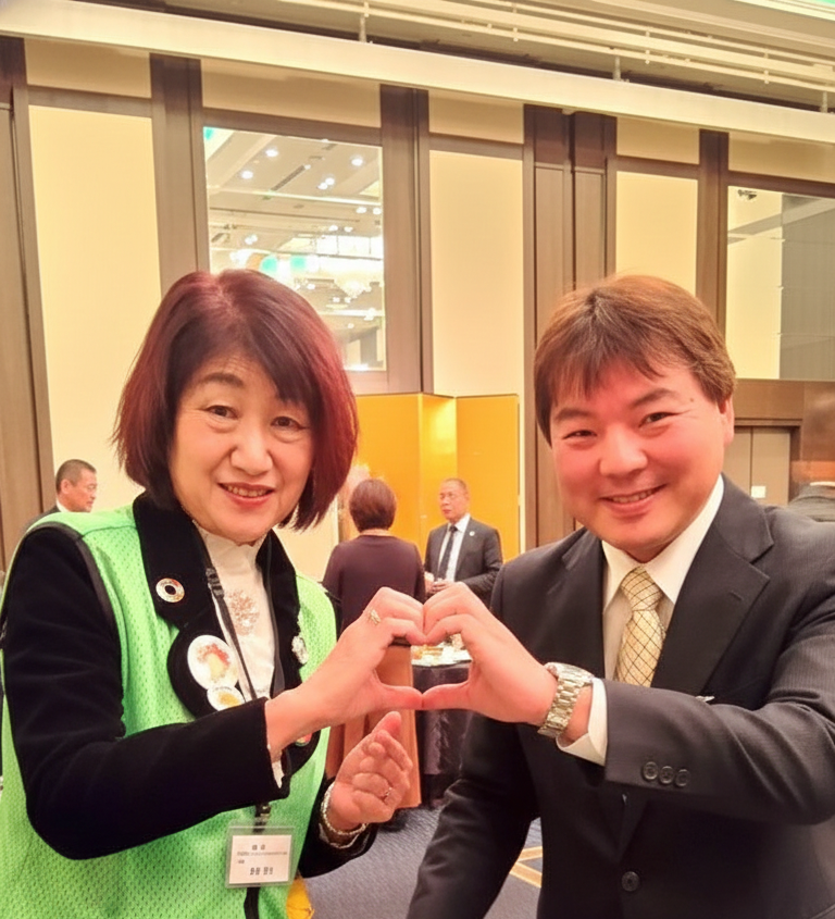 懇親会会場での野村代表（左）と投稿者（右）のツーショット。野村氏はひまわりがデザインされた緑の活動ベストを着用し、二人は笑顔で飲み物や皿を手にしている。