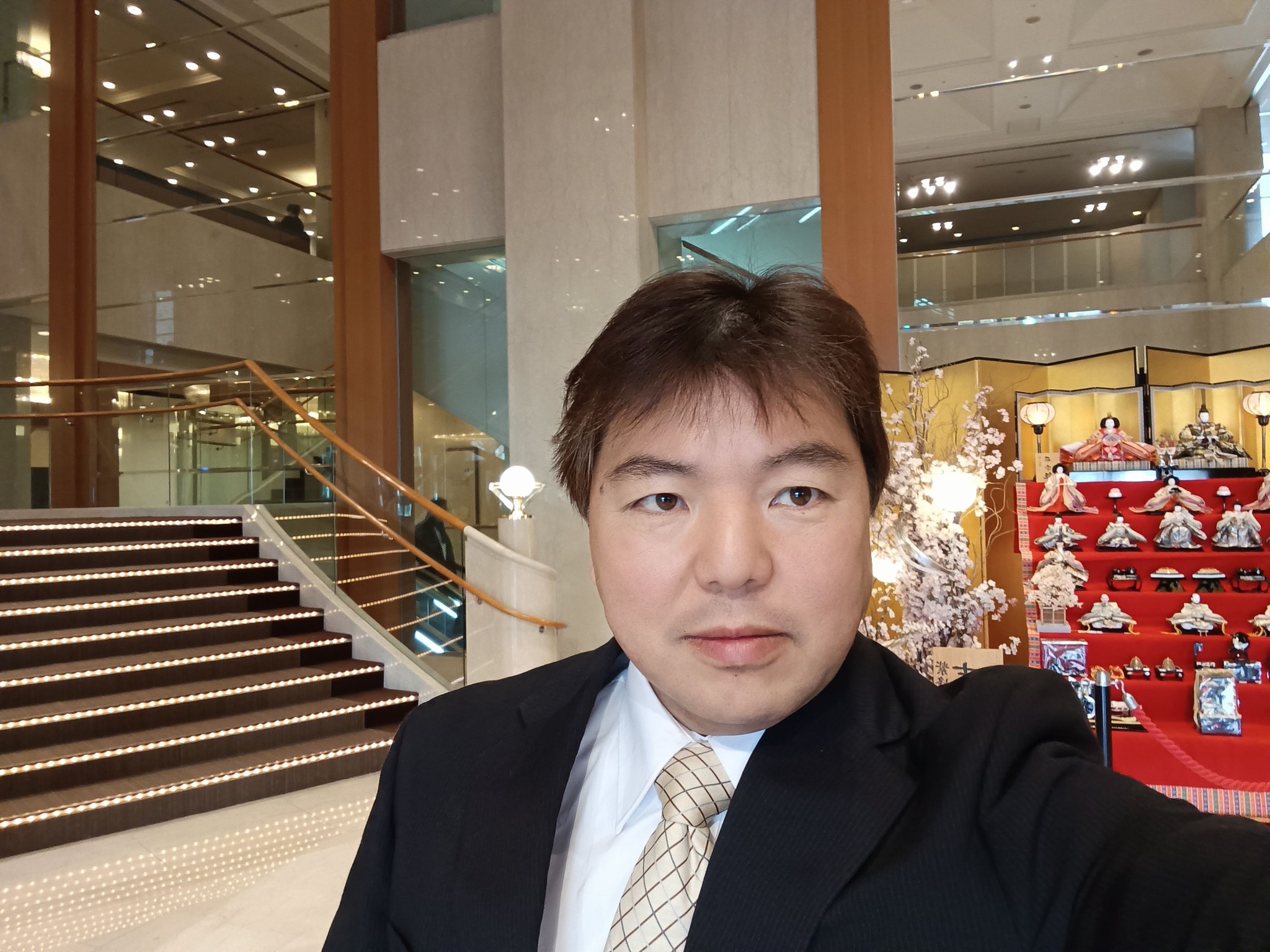ロビーの雛人形を背景に、スーツ姿の榊原平が微笑む自撮り写真。