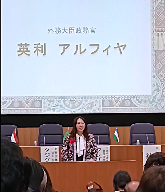 シンポジウムの壇上でスピーチをする英利アルフィヤ外務大臣政務官。背後のスクリーンには「外務大臣政務官 英利 アルフィヤ」の文字と、中央アジアの伝統的な織物風の模様が映し出されている。壇上には中央アジア各国の卓上旗が並び、政務官は華やかな柄のジャケットに赤いコサージュを身につけている。