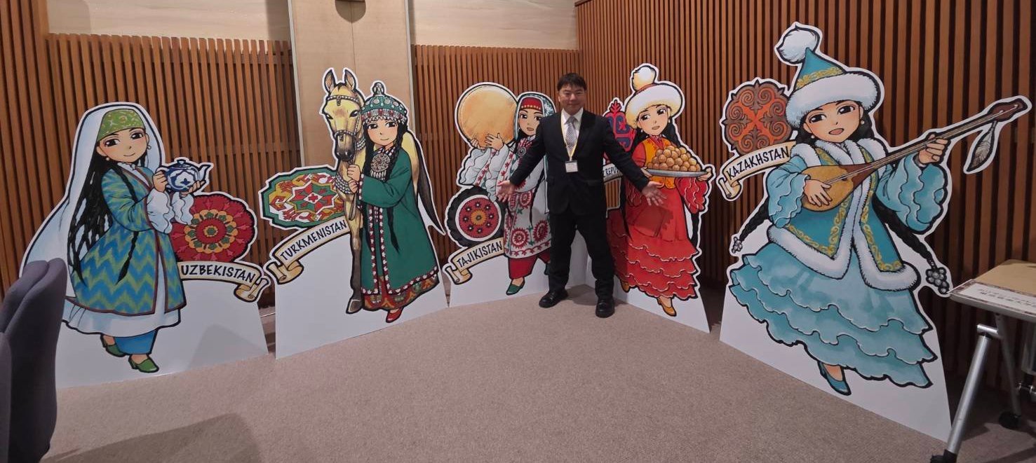 中央アジア5か国の鮮やかな民族衣装を纏った女性キャラクターたちの等身大パネル。その中央に立ち、両手を広げてシンポジウムの活気ある雰囲気を伝えるようにポーズをとるスーツ姿の榊原平。