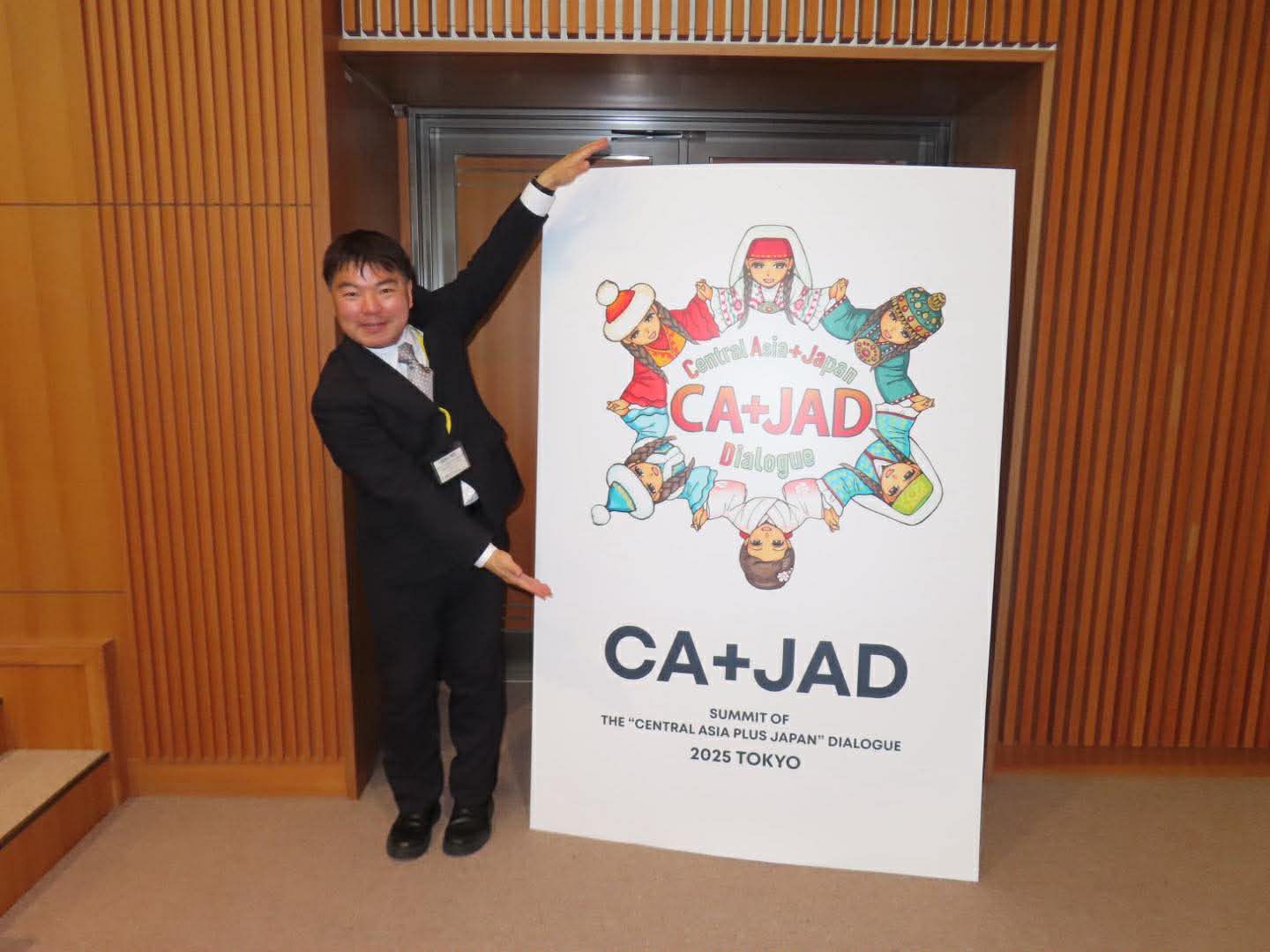 「CA+JAD」の大型ポスターの横で、左手を高々と掲げてポーズをとる榊原平。ポスターには中央アジアのキャラクターたちが描かれ、イベントへの参加を記念した一枚。